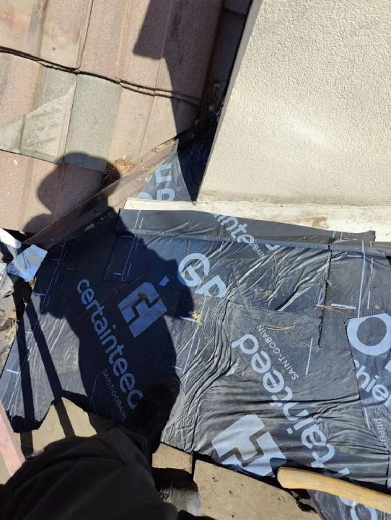 A person stands on a roof near tiles and a wall, with CertainTeed and GP underlayment visible beneath their feet. Shadows are cast across the work area.