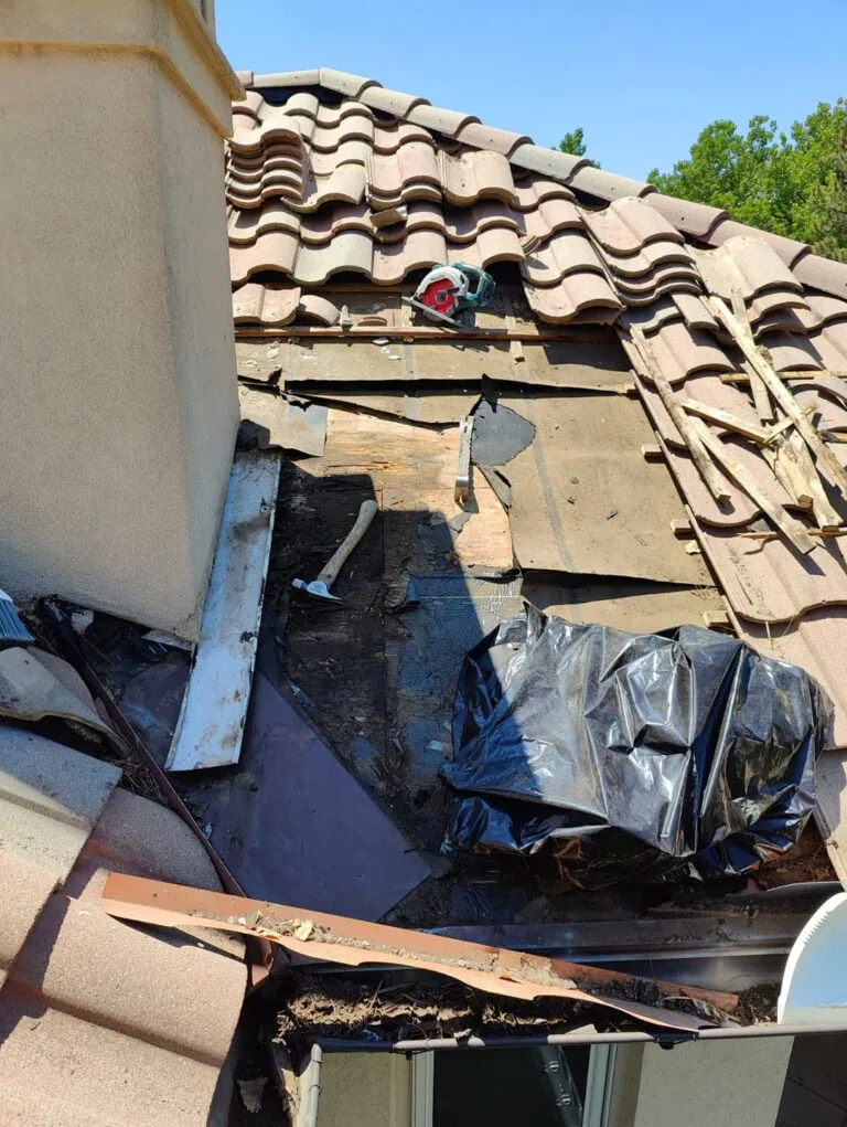A damaged tile roof next to a chimney, with tiles removed, debris, tools, and a black plastic sheet covering part of the exposed area.