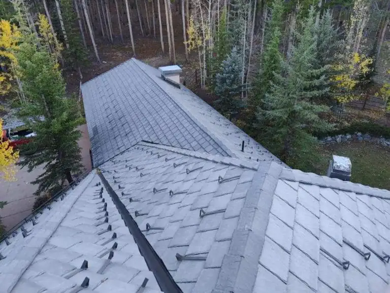 Close-up of a tiled roof with curved, overlapping shingles and a large white chimney. The tiles are grayish-brown and some have visible seams and shadows from the sunlight.