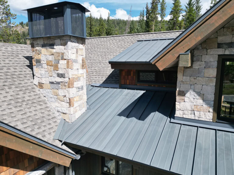 A close-up view of a house roof shows gray metal and shingle sections, a stone chimney with a metal cap, and surrounding trees in the background under a partly cloudy sky.
