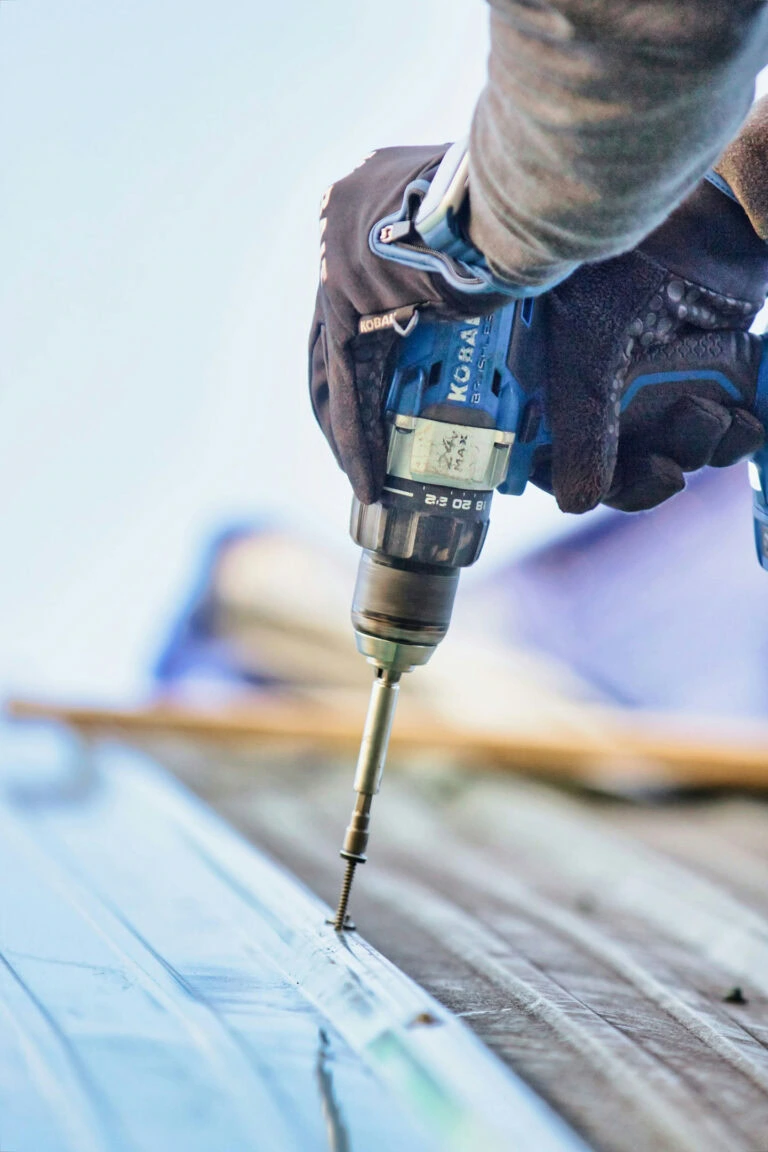 A person wearing gloves uses a cordless power drill to drive a screw into a metal surface, likely working on construction or repairs outdoors.