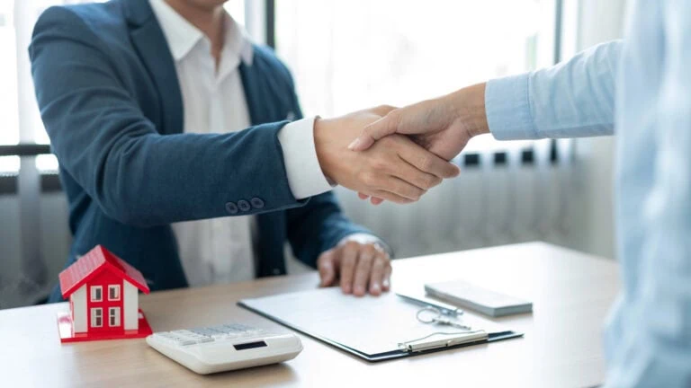 Two people shaking hands across a desk with a small red house model, calculator, clipboard, and documents, suggesting a real estate or business agreement and addressing any misconception on roof warranty.