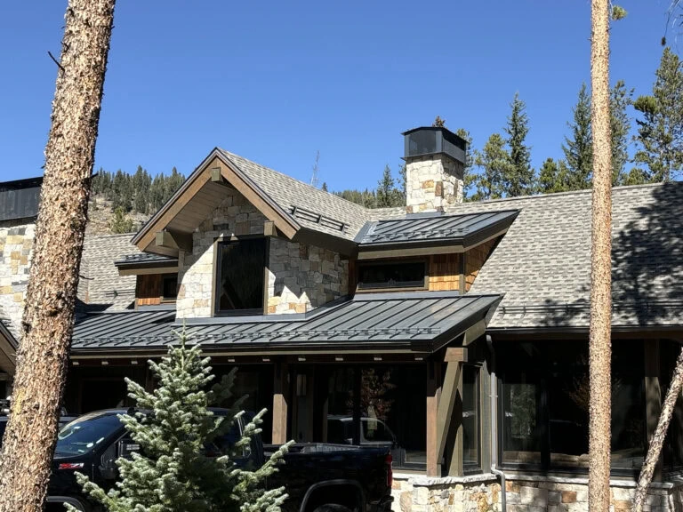 A modern house with stone and wood siding, metal roof accents, large chimney, and multiple peaked gables sits among tall pine trees under a clear blue sky. A black truck is parked in front.